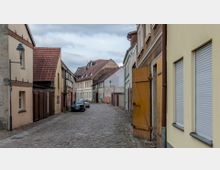 Eine enge gepflasterte Gasse in einer Altstadt mit historischen Gebäuden. Die Umgebung ist geprägt von Fachwerkhäusern, gelb- und beigefarbenen Fassaden sowie einem offenen Holztor auf der rechten Seite.