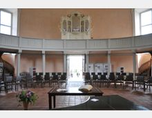 Innenraum einer schlichten Kirche mit halbkreisförmiger Anordnung von Stühlen, einer Empore mit weiß-goldener Orgel im Hintergrund und einer geöffneten Doppeltür, durch die Tageslicht einfällt. Im Vordergrund steht ein Tisch mit offenem Buch und einem Blumenstrauß.