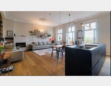 Helles Wohnzimmer mit offener Küche, großen Fenstern und Zugang zu einem Balkon. Der Raum enthält eine große graue Couch, ein modernes schwarzes Küchenmodul mit Spüle, hölzerne Esstischmöbel und dekorative Regale an der Wand.