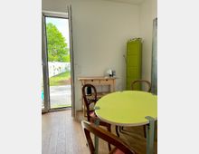 Ein heller Raum mit Holzboden, ausgestattet mit einem runden, hellgrünen Tisch und holzgerahmten Stühlen. Ein großes Glastürfenster führt zu einem Gartenbereich mit Rasen und gepflastertem Boden, während im Hintergrund ein grüner Metallspind und ein Holztisch mit Geräten stehen.