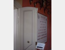 Weiß lackierte Tür in einem historischen Gebäude, daneben eine Wand mit einem Stammbaum-Diagramm und Wappensymbolen. Darüber befinden sich an der roten Wand Tiergeweihe, und im Vordergrund steht ein Schild mit der Aufschrift „Rundgang und zur Kasse“.