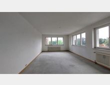 Leerer, rechteckiger Raum mit weißen Wänden und einer Reihe von Fenstern auf zwei Seiten, die Blick auf Bäume bieten. Der Boden ist aus Beton, und an den Wänden befinden sich Heizkörper unter den Fenstern.