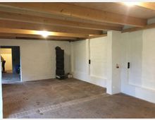 Ein leerer Raum mit weißen, verputzten Wänden, einer Decke mit sichtbaren Holzträgern und einem Boden aus roten Ziegelsteinen. Im Hintergrund ist eine geöffnete Tür zu einem angrenzenden Raum sichtbar, und an der rechten Wand steht ein kleiner schwarzer Kamin.