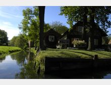 Ein rustikales Ferienhaus mit Fachwerkdetails steht neben einem kleinen, bewachsenen Kanal, umgeben von Bäumen und Grasflächen; ein Schild mit Vermietungsinformationen ist im Vordergrund sichtbar.