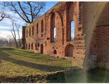 Ruine einer mittelalterlichen Backsteinkirche mit erhaltenen gotischen Spitzbögen und großen, offenen Fensteröffnungen; umgeben von einer grasbewachsenen Fläche und kahlen Bäumen bei klarem Himmel.