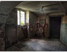 Ein verlassener Innenraum mit abblätternden Wänden und einer vergilbten Decke aus Beton. Ein kleines Fenster lässt Tageslicht herein, während eine offene Tür und ein Aushang an der Wand sichtbar sind; das Umfeld wirkt heruntergekommen.