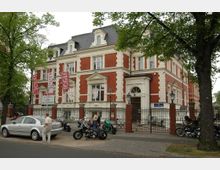 Ein freistehendes, mehrstöckiges Geschäftshaus im neoklassizistischen Stil mit roten Ziegelwänden und weißen Putzverzierungen. Vor dem Gebäude befindet sich ein Zaun mit Tor, parkende Motorräder und ein Auto sowie vereinzelte Passanten; im Vordergrund stehen große Bäume.