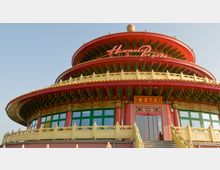 Großes, mehrstöckiges Gebäude im Stil einer traditionellen chinesischen Pagode, mit roten, runden Dächern und goldenen Verzierungen. Der Eingang ist mit grünen Fenstern, roten Säulen und einer Treppe mit gelbem Geländer gestaltet.