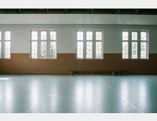 Innenansicht einer Turnhalle mit großen, halbkreisförmig abgeschlossenen Fenstern, weiß gestrichenen Wänden und einer hellgrauen Spielfeldfläche aus Kunststoffboden. Die untere Wandhälfte ist braun verkleidet, und im Hintergrund stehen einfache Holzbänke.