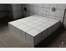 Plattform aus weißen Fliesen in einem Raum mit gefliesten Wänden und dunklem Boden. An der Wand befinden sich eine Steckdose und Rohröffnungen.