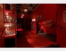 Ein schmaler, rot beleuchteter Innenraum mit roten Wänden und Teppichboden. Links befindet sich eine Glasvitrine mit dekorativen Objekten, rechts eine Garderobe mit Kleiderbügeln, und im Hintergrund ein kleiner Empfangstresen unter mehreren Kronleuchtern.