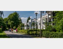 Moderner Wohnkomplex mit weißen Gebäuden und Balkonen, umgeben von Bäumen und gepflegten Gärten. Im Vordergrund führt ein gepflasterter Weg entlang, auf dem mehrere Personen mit Fahrrädern unterwegs sind; Laternen und Grünflächen säumen den Weg.