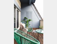 Eine moderne Dachterrasse mit Holzboden, grünen Stühlen aus Seilmaterial, einem gläsernen Beistelltisch und dekorativen Pflanzen. Im Hintergrund befindet sich eine blaue Holzverkleidung mit Fensterläden.