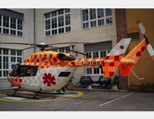 Ein orange-weiß gemusterter Rettungshubschrauber mit der Aufschrift „112“ und einem roten Sternsymbol steht auf einer markierten Landeplattform in einem Innenhof, umgeben von hellen Ziegelgebäuden mit großen Fenstern.