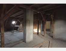 Ein leerer Dachboden mit Holzbalken und Holzboden, teilweise beschädigt, sowie einer kleinen Dachgaube, die Licht hineinlässt. Im Raum sind ein massiver Betonpfeiler und offene Wand- und Türbereiche sichtbar.