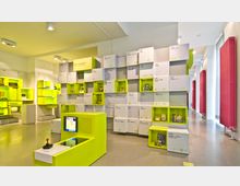 Ein modernes Ausstellungsdesign in einem Museum, mit gelb-grün leuchtenden Regalen, die verschiedene Exponate und Informationstafeln zu Computern und Videospielen präsentieren. Der Raum ist hell beleuchtet, mit schlichtem Boden und hohen Fenstern, die teils rotfarbene Heizkörper zeigen.