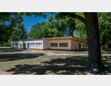 Ein eingeschossiges, weißes Gebäude mit Flachdach und überdachtem Eingangsbereich, umgeben von einer parkähnlichen Grünfläche mit großen Bäumen. Die Fenster des Gebäudes sind mit Rollläden verschlossen, und der Boden besteht aus trockenem Gras und Erde.