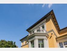 Außenansicht eines mehrstöckigen historischen Wohnhauses mit gelblicher Fassade, weißen Gesimsen und Erkern sowie einem kleinen Balkon mit Metallgeländer. Im Hintergrund sind blauer Himmel und Baumkronen zu sehen.
