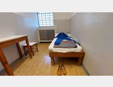 Schlichte Gefängniszelle mit einem Holzbett, auf dem eine blaue Gefängnisuniform und eine zusammengefaltete Decke liegen. Links steht ein kleiner Holztisch mit Hocker, das Fenster hat Gitterglas, und der Fußboden besteht aus hellem Holz.