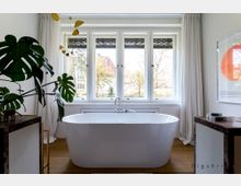 Modernes Badezimmer mit freistehender, weißer Badewanne vor einer großen Fensterfront mit weißen Vorhängen. Links im Bild eine große Monstera-Pflanze, rechts ein Kunstbild mit einem orangefarbenen Kreis, Boden aus Holz.