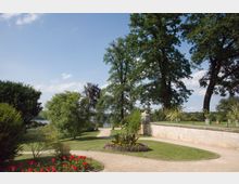 Ein gepflegter Park mit geschwungenen Kieswegen, blühenden Blumenbeeten, grünen Rasenflächen und großen Bäumen. Im Hintergrund ist eine niedrige Steinmauer sowie ein See sichtbar, umgeben von dichter Vegetation und blauen Himmel.