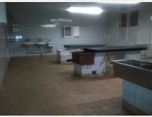 Ein einfacher, gefliester Raum mit Fliesenwänden und braunem Bodenbelag, ausgestattet mit zwei Waschbecken, mehreren Arbeitstischen und einem großen Spülbecken; typisch für eine Arbeitsküche oder einen Funktionsraum. Ein Fenster und eine helle Deckenleuchte sind ebenfalls sichtbar.