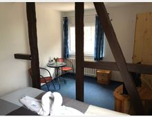 Hotelzimmer mit Holzbalkenelementen, einem Bett im Vordergrund, einem kleinen runden Tisch mit zwei Stühlen und einem Fenster mit blauen Vorhängen im Hintergrund. Der Raum hat einen blauen Teppichboden, Holzmöbel und wirkt schlicht eingerichtet.