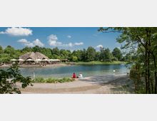 Ein See mit klarer Wasseroberfläche, umgeben von Bäumen und Grünflächen. Auf der linken Seite befindet sich ein caféartiges Gebäude mit Zeltdächern und Sitzbereichen am Ufer, während im Vordergrund Personen auf einer gepflasterten Fläche sitzen.