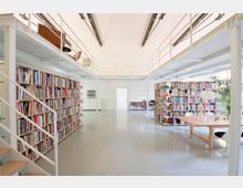 Ein moderner, heller Bibliotheksraum mit hohen Decken, offenen Regalen voller Bücher, einer Galerieebene mit Geländer und einer Treppe. Im Vordergrund steht ein großer Holztisch, und Pflanzen sind dezent in die Einrichtung integriert.