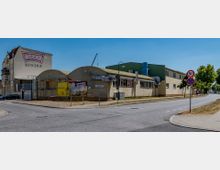 Eckansicht einer Fabrik mit gelblichen Gebäuden, einem großen "Werder Ketchup"-Schriftzug auf der Fassade eines angrenzenden Hauses und Werbeplakaten entlang der Straße. Die Szene zeigt eine asphaltierte Kreuzung mit Straßenschildern und Gehwegen, umgeben von vereinzelten Bäumen.