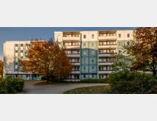 Wohnblock mit fünf Stockwerken in heller und pastellgrüner Farbgestaltung, umgeben von Gehwegen, Bäumen in Herbstfarben und Buschwerk. Im Vordergrund führt ein geschwungener Weg durch eine kleine Grünanlage.