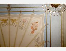 Decke eines prunkvollen Raumes mit dekorativen Malereien, darunter ein Pegasus, florale Motive und eine Frauenfigur in einem Oval. Goldene Zierelemente, hellblaue und cremefarbene Flächen sowie ein filigraner Kronleuchter betonen das aufwendige Design.
