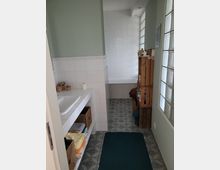 Ein Badezimmer mit hellgrünen Wänden und einer weißen Kachelausstattung. Zu sehen sind ein Waschbecken mit weißer Fliesenumrandung, Regalfächer mit Körben, eine geflieste Badewanne im Hintergrund und ein Holzkastenregal rechts neben der Badewanne. Der Boden ist mit gemusterten Fliesen ausgelegt, und eine Glasbausteinwand lässt Tageslicht herein.