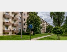 Mehrstöckige Wohngebäude mit beige- und braunfarbenen Fassaden und Balkonen, umgeben von einer Grünanlage mit Bäumen, Rasen und einem Gehweg. Im Vordergrund steht ein Verkehrsschild, das einen gemeinsamen Fußgänger- und Radweg anzeigt.