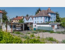 Wohngebiet mit kleinen Gärten und Gebäuden: Im Vordergrund ein bepflanzter Gartenbereich mit Wegen, ein kleines weißes Gartenhaus mit einer Bank unter einer Überdachung. Im Hintergrund mehrstöckige Wohnhäuser in blauer und cremefarbener Fassade, umgeben von Bäumen und Hecken.