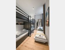 Schmales, modern eingerichtetes Schlafzimmer mit schwarzen Metall-Etagenbetten auf der linken Seite und einem Einzelbett am Fenster rechts. Helle Holzböden, weiße Wände, Einbauspots an der Decke sowie ein Spiegel und ein Vorhang am Ende des Raums.