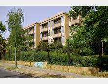 Mehrstöckiges Wohngebäude mit gelblichen Klinkerfassaden, breiten Balkonen und großen Fenstern, umgeben von dichter Vegetation. Im Vordergrund eine niedrige Ziegelmauer mit schwarzem Metallzaun sowie Gehweg und Straßenlaterne.