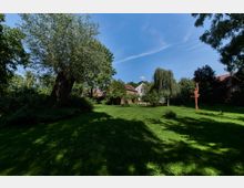 Grüne Parklandschaft mit einer großen Rasenfläche, umgeben von Bäumen und Büschen. Im Hintergrund sind Gebäude mit roten und weißen Dächern sichtbar, daneben eine Holzskulptur und eine Parkbank unter einem hängenden Baum.