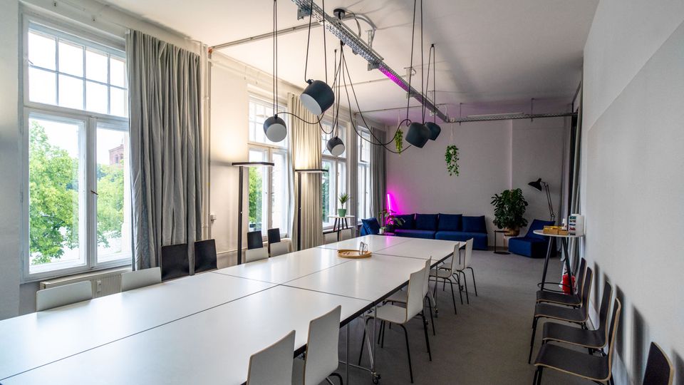Heller Konferenz- und Arbeitsraum mit langen weißen Tischen und Stühlen, hohen Fenstern mit grauen Vorhängen und mehreren hängenden Deckenlampen. Am hinteren Ende stehen blaue Sofas neben Pflanzen, ergänzt durch eine pinke Akzentbeleuchtung.