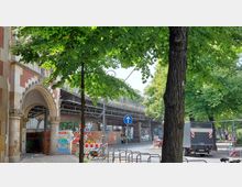 Ein urbaner Straßenzug mit einem gemauerten Torbogen an einem Gebäude aus gelben und roten Backsteinen im Vordergrund. Im Hintergrund verläuft eine erhöhte Bahntrasse, umgeben von grünen Bäumen, geparkten Autos und Baustellenabsperrungen.