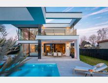 Modernes, zweigeschossiges Wohnhaus mit klaren, kubischen Formen, großen Glasfronten und überdachter Terrasse mit Esstisch. Davor liegen eine gepflasterte Poolterrasse mit rechteckigem Swimmingpool und Liege, umgeben von Rasenfläche und dunklem Zaun.