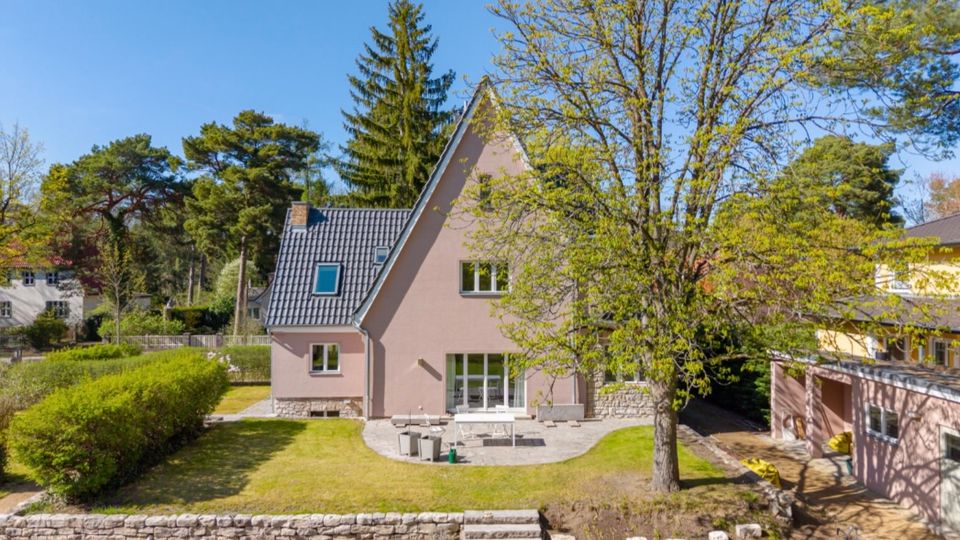 Freistehendes Einfamilienhaus mit steilem Satteldach und hellrosa Fassade, davor ein gepflegter Garten mit Terrasse, Rasenfläche, niedriger Steinmauer und Treppenstufen. Umgeben von hohen Bäumen und Hecken in einer ruhigen Wohngegend bei blauem Himmel.
