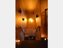 Treppe in einem schummrig beleuchteten, engen Innenraum mit warmen, goldbraunen Wänden. Wandleuchten und dekorative Kerzen erhellen den Bereich, während schwarze Dekorationen in Form von Kaninchenköpfen an den Wänden angebracht sind.