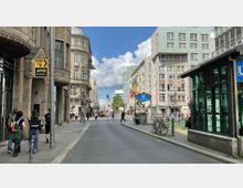 Straßenszene mit historischen und modernen Gebäuden in Berlin, nahe Checkpoint Charlie. Im Vordergrund sind Straßenschilder, Fahrräder und ein U-Bahn-Zugang der Station "Kochstraße" zu sehen, während Passanten die Straße überqueren.