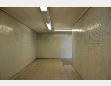 Ein leerer, rechteckiger Tunnel mit kahlen Wänden aus kleinen, hellen Fliesen. Der Boden besteht aus großformatigen, grauen Fliesen, und an der Decke sind zwei längliche Leuchtstofflampen angebracht.