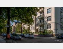 Straßenszene in einem Wohngebiet mit mehrstöckigen Gebäuden im hellen Fassadenstil und Balkonen, umgeben von Bäumen und gepflegten Vorgärten. Geparkte Autos säumen die Straße, und Straßenschilder markieren die Kreuzung der Varziner Straße und der Schmiljanstraße.