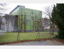 Ein modernes Gebäude mit einer grünen Außenfassade, teilweise von rankenden Pflanzen bewachsen, umgeben von einem Maschendrahtzaun. Im Vordergrund liegt eine Rasenfläche, im Hintergrund sind kahle Bäume und weitere Gebäude sichtbar.