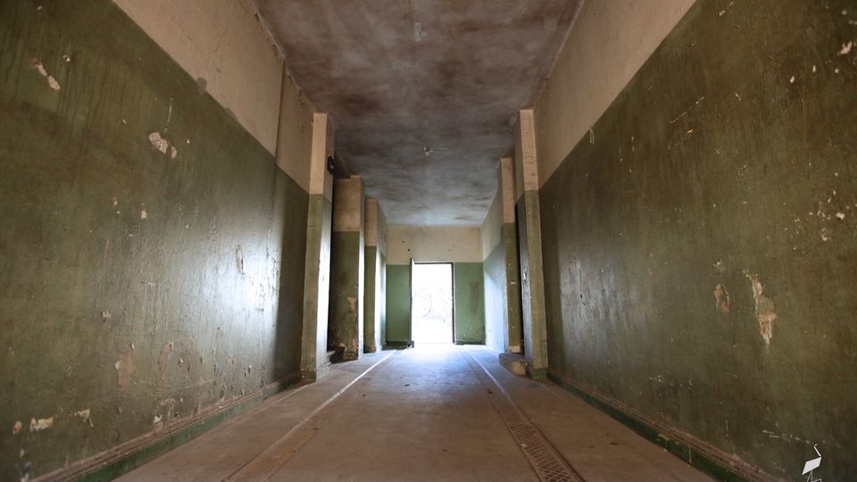 Langer, schmaler Innenkorridor mit abgenutzten grün-beigen Wänden, Betonboden mit eingelassenen Rinnen und einer niedrigen Decke. Mehrere Stützpfeiler gliedern den Raum, am Ende führt eine offene Tür in einen hellen Außenbereich.