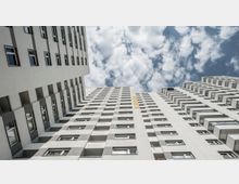 Blick nach oben auf mehrere hohe, moderne Wohngebäude mit weißen Fassaden und zahlreichen Fenstern sowie Balkonen. Im Hintergrund ist ein Himmel mit vereinzelten Wolken zu sehen.