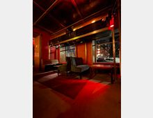 Innenraum einer Bar oder Lounge mit roten Wänden, gemustertem Teppich und dunklen Sitzbänken sowie Sesseln um niedrige Tische. Über den Fenstern verläuft eine goldfarbene Metallkonstruktion unter einer verzierten Kassettendecke, mit warmem, rötlichem Licht und Blick nach draußen auf die Straße.